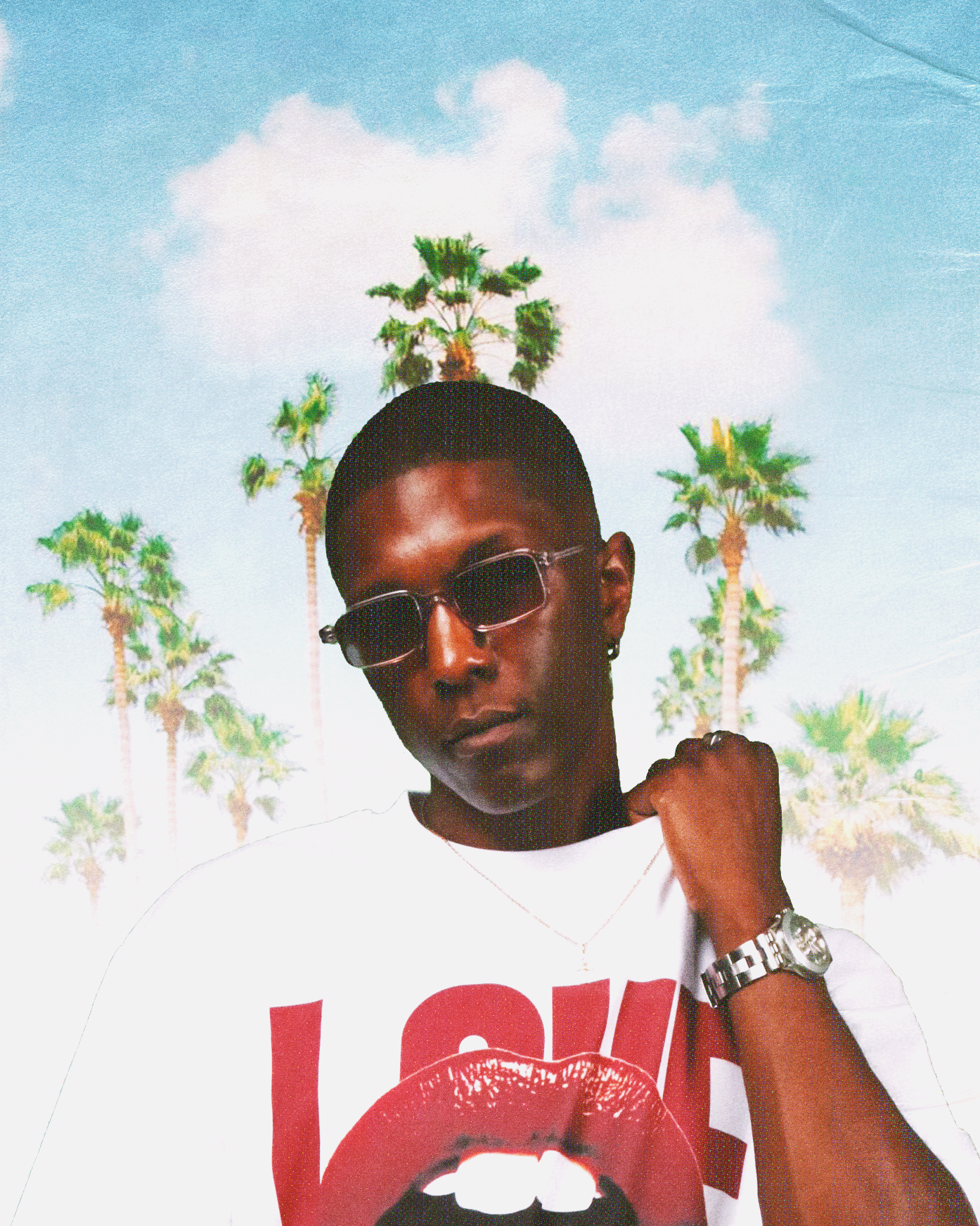 A fashion-forward black man, holding a T-shirt by the collar with a silver watch on his wrist and wearing a bright red graphic T-shirt adorned with red lips, poses in front of tropical palm trees and a blue sky—a trendy summer streetwear style and a vacation vibe.