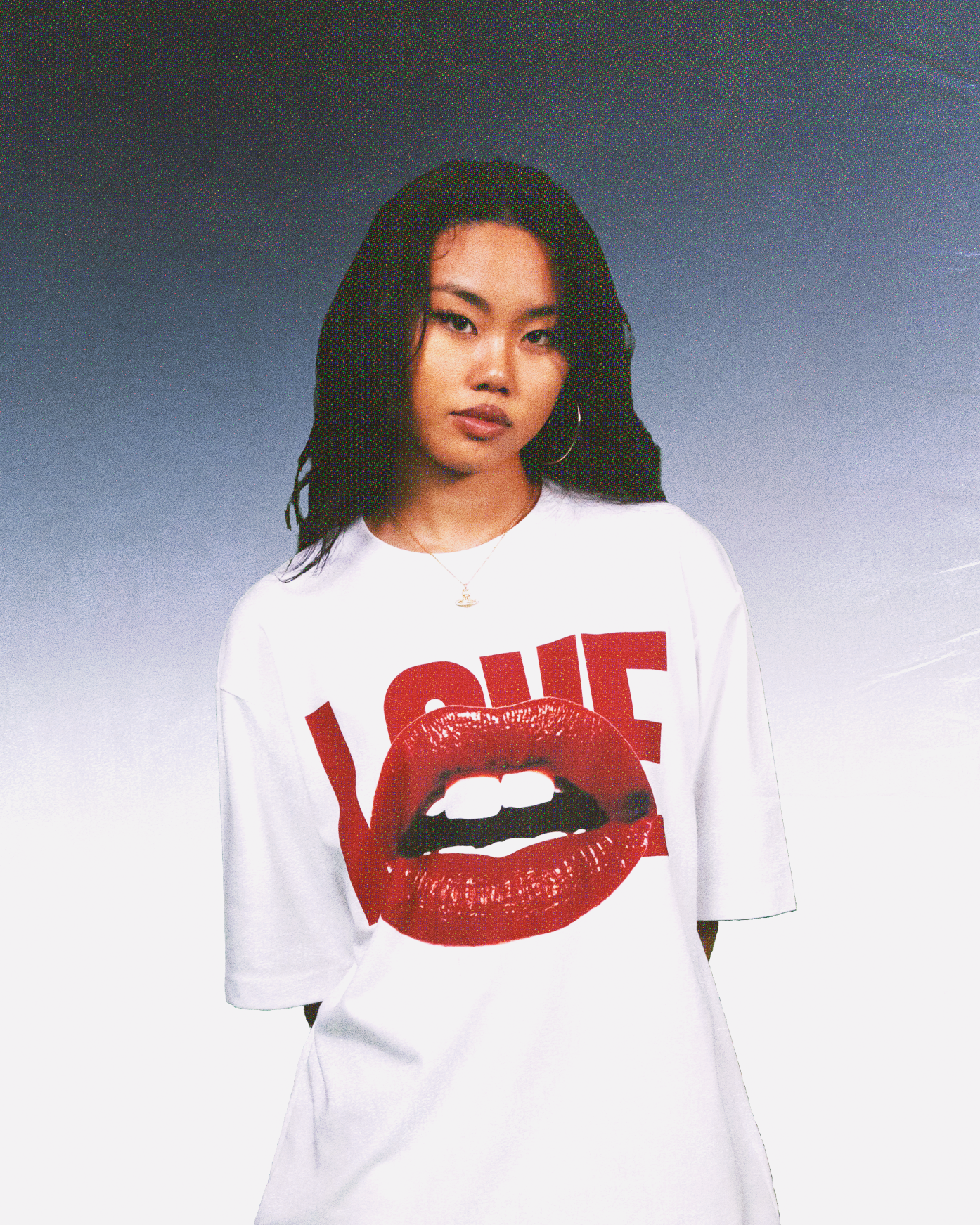 Young woman with long black hair wearing a white oversized T-shirt featuring bold red 'LOVE' text and glossy red lips graphic, standing against a gradient blue-to-white background.