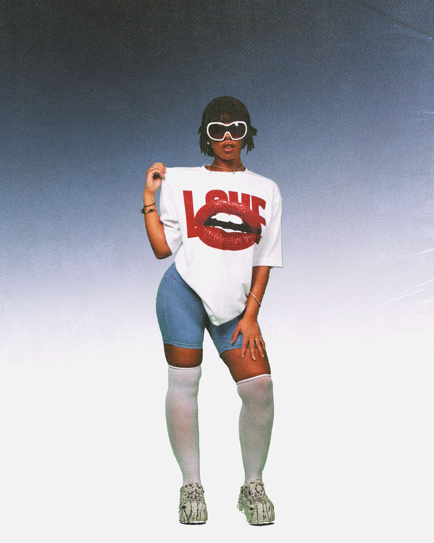 Stylish woman wearing oversized white graphic T-shirt with bold red 'LOVE' text and glossy lips print, paired with blue cycling shorts, white thigh-high socks, chunky patterned sneakers, and oversized white sunglasses, posing against a gradient blue-to-white background.